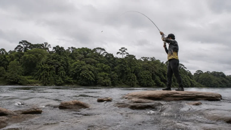 Pescador lançando a linha com carretilha em cima de uma pedra à beira de um rio de água doce