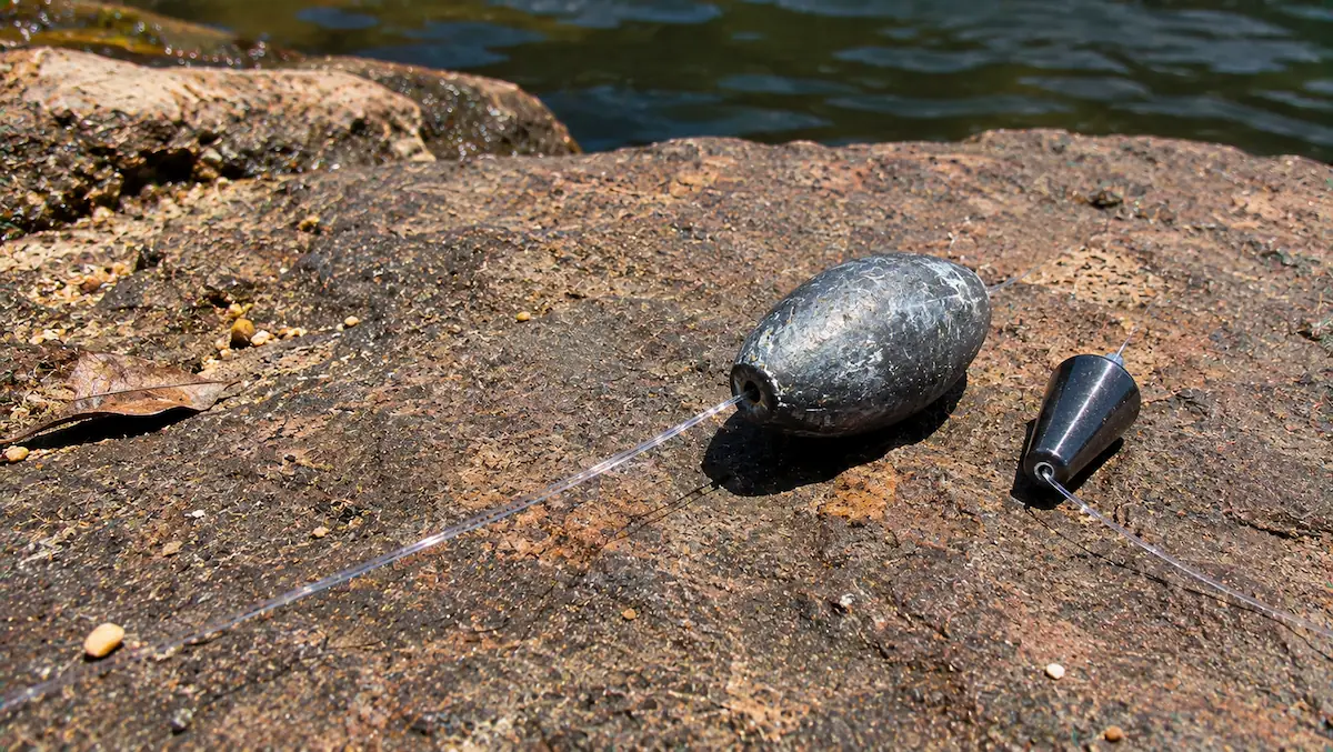Chumbada oliva de chumbo fosca ao lado de um peso de tungstênio menor e escuro, com o mesmo peso, sobre uma pedra de rio